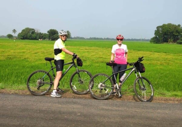 Two cyclists in colorful attire pause with their bikes on a rural road, surrounded by green fields and distant trees under a clear sky.