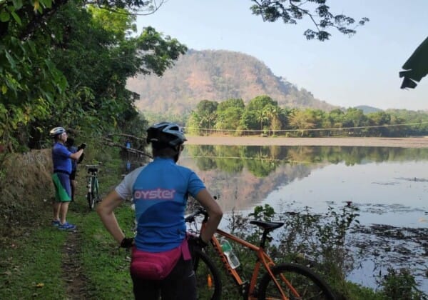 Two cyclists riding their bicycles on a path next to a serene lake surrounded by lush green trees and mountains in the background.