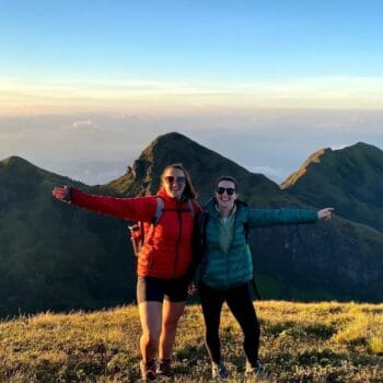 Two women smile for a photo at the summit of Meshapulimala, surrounded by stunning mountain views.