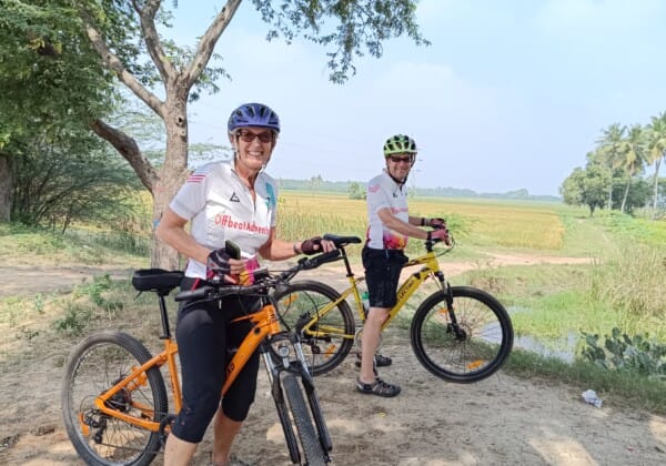 Two people on bikes stand side by side, smiling and enjoying a sunny day outdoors.